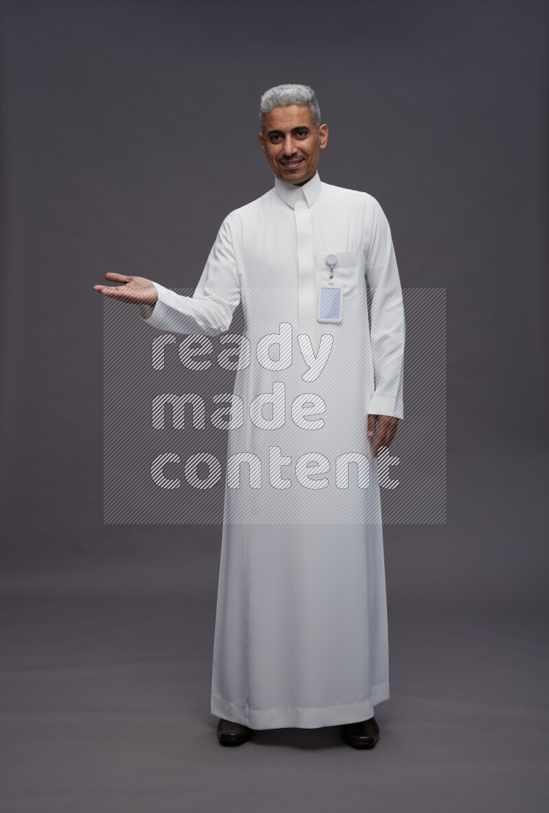 Saudi man wearing thob with pocket employee badge standing interacting with the camera on gray background