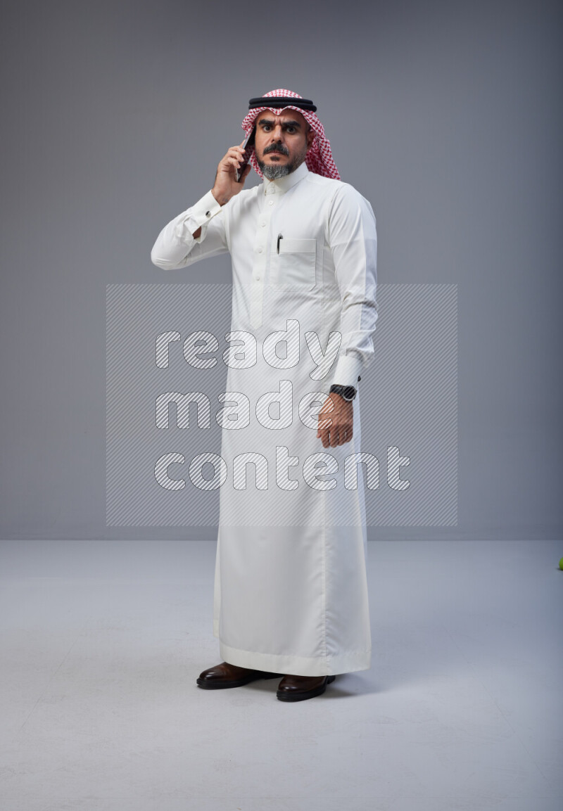 Saudi man Wearing Thob and red Shomag standing talking on phone on Gray background