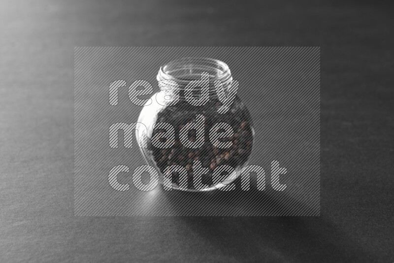 A glass spice jar full of black peppers on black flooring