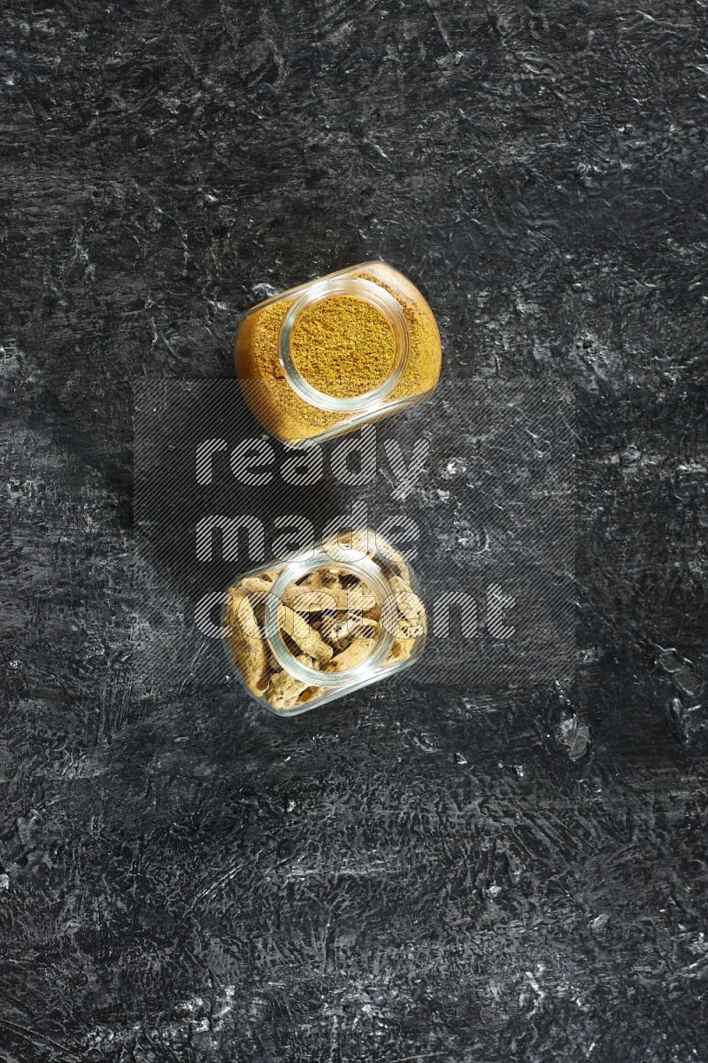 وعاءان زجاجيان للتوابل ممتلئان ببودرة الكركم وأصابع الكركم المجففة على خلفية سوداء