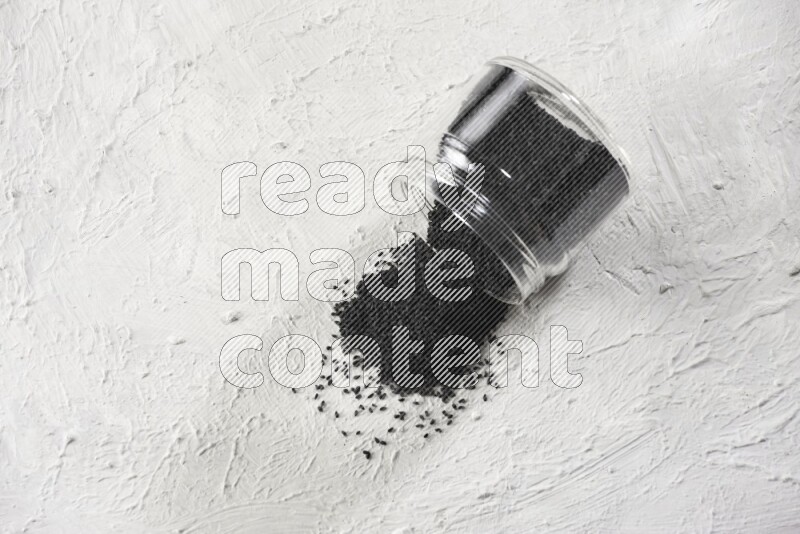 A glass jar full of black seeds and the jar flipped and seeds spread on a textured white flooring