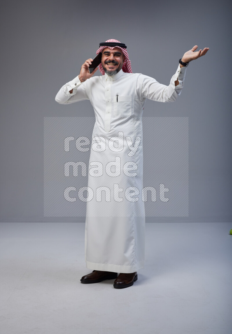 Saudi man Wearing Thob and red Shomag standing talking on phone on Gray background