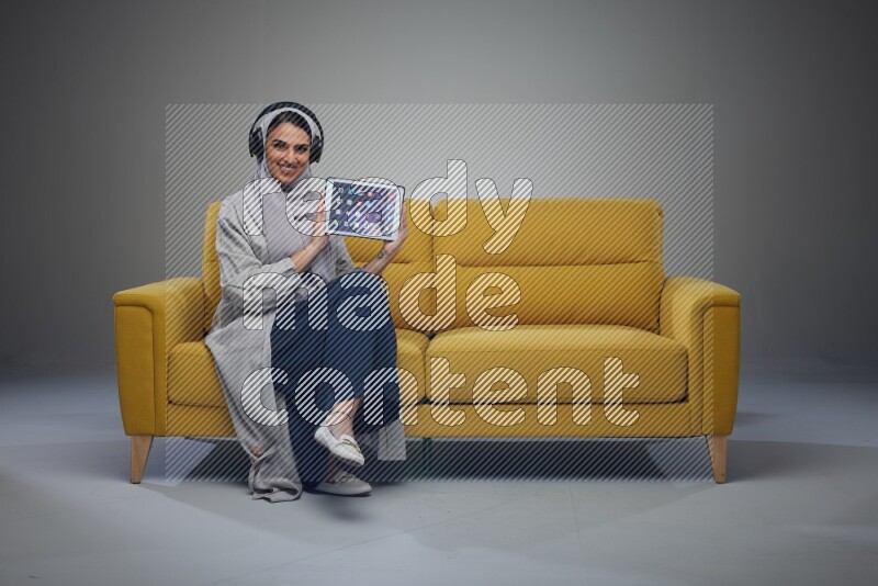 A Saudi female wearing a light gray Abaya and head scarf sitting on a yellow sofa and showing her tablet's screen while wearing headphones eye level on a grey background