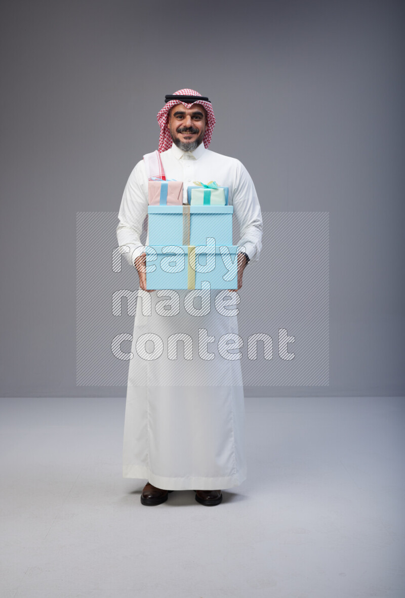 Saudi man Wearing Thob and red Shomag standing holding gift box on Gray background