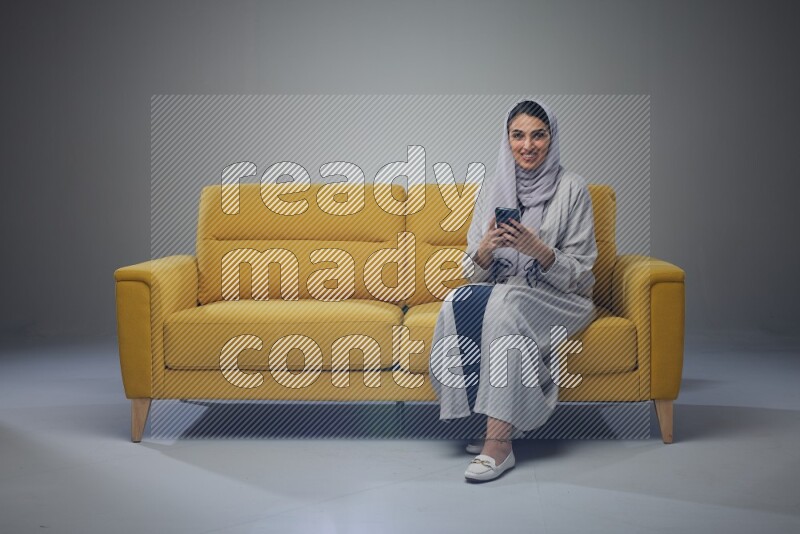 A Saudi woman wearing a light gray Abaya and white head scarf sitting on a yellow sofa and using her phone eye level on a grey background