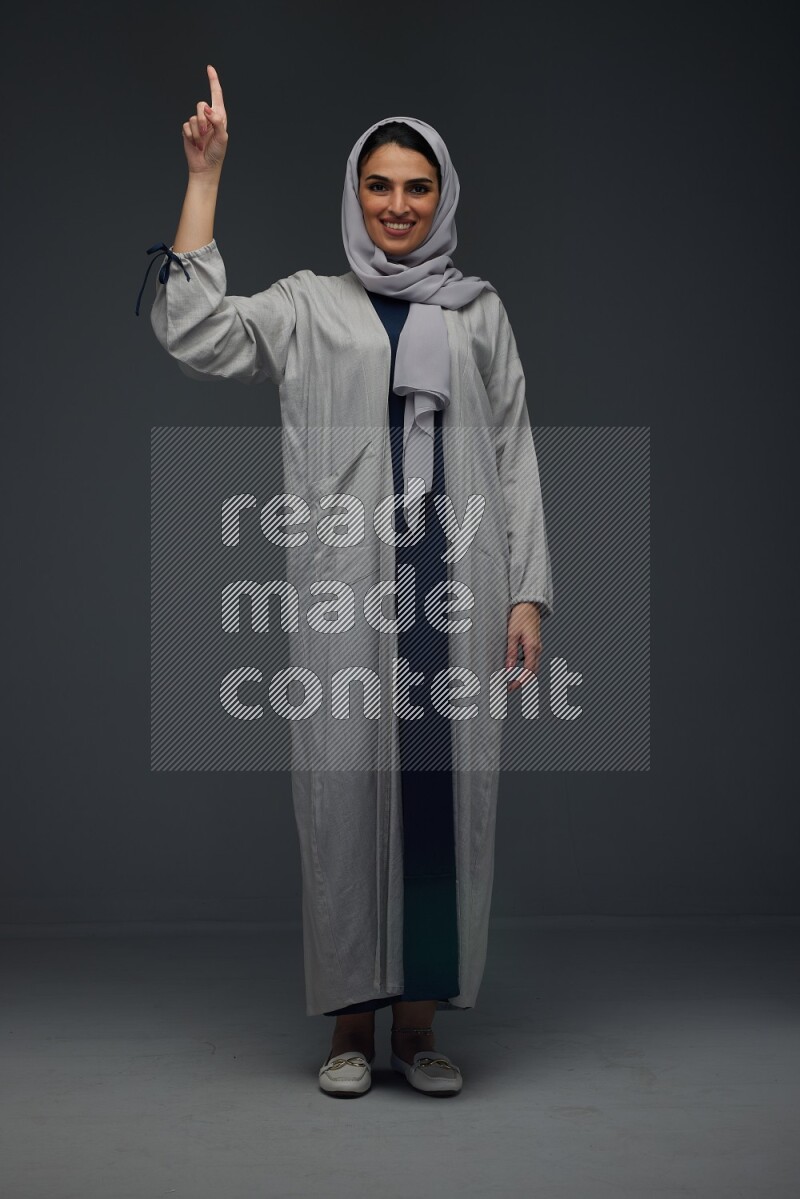 A Saudi woman wearing a light gray Abaya and head scarf standing and making multi poses eye level on a grey background