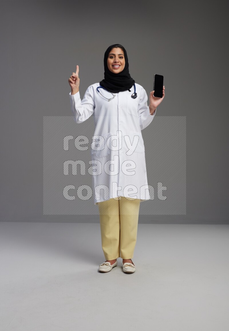 Saudi woman wearing lab coat with stethoscope standing showing phone to camera with sign in the back on Gray background