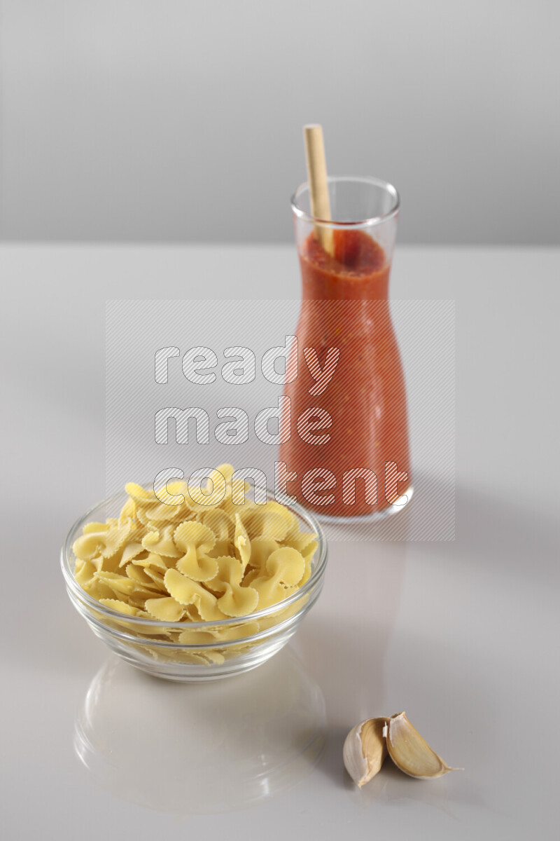 Raw pasta with tomatoe pasta with different ingredients such as cherry tomatoes, basil, garlic, bay laurel, cardamom, white pepper, black pepper, red chilis and wheat stalks on light grey background