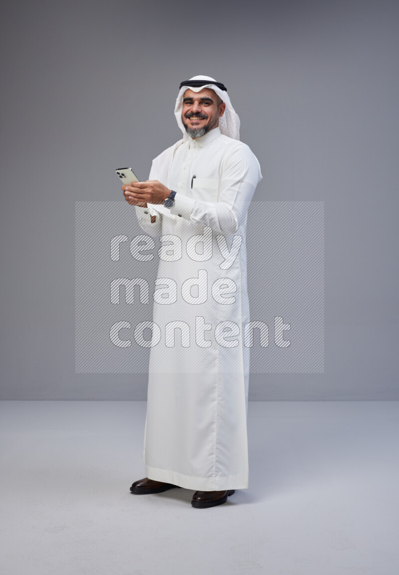 Saudi man Wearing Thob and white Shomag standing texting on phone on Gray background