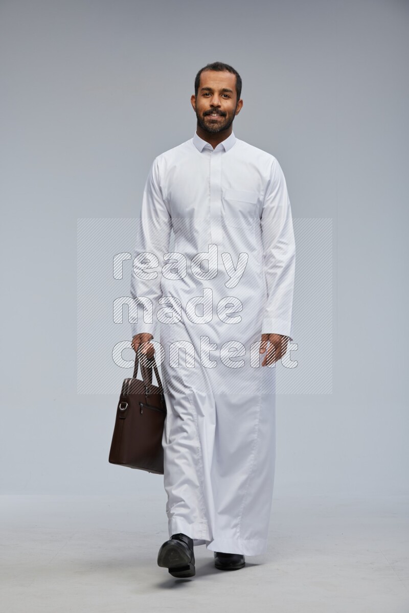 Saudi man wearing thob standing holding suitcase on gray background