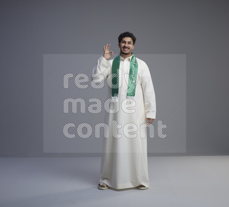 A Saudi man standing wearing thob and Saudi flag scarf on gray background
