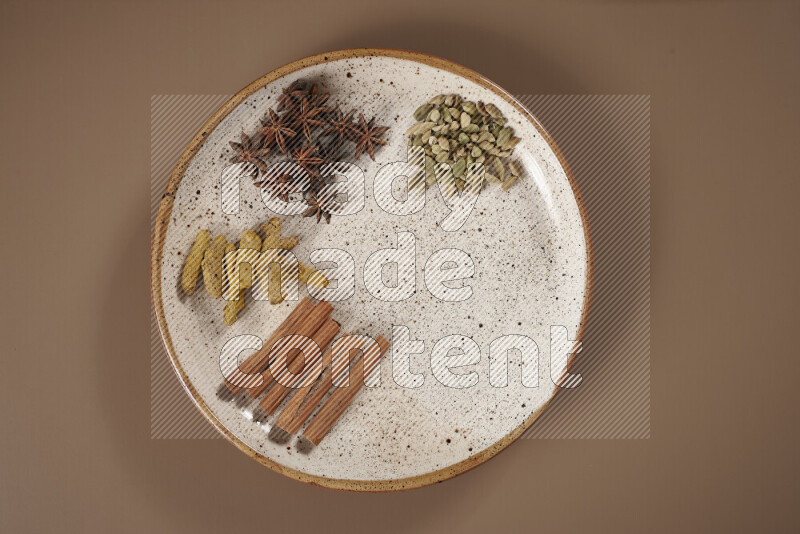 An assorted whole spices on a flat beige plate including fresh and dried ginger, cinnamon sticks, cardamom, turmeric fingers, cloves, star anise on a beige background
