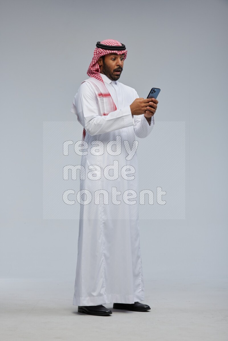Saudi man Wearing Thob and shomag standing texting on phone on Gray background