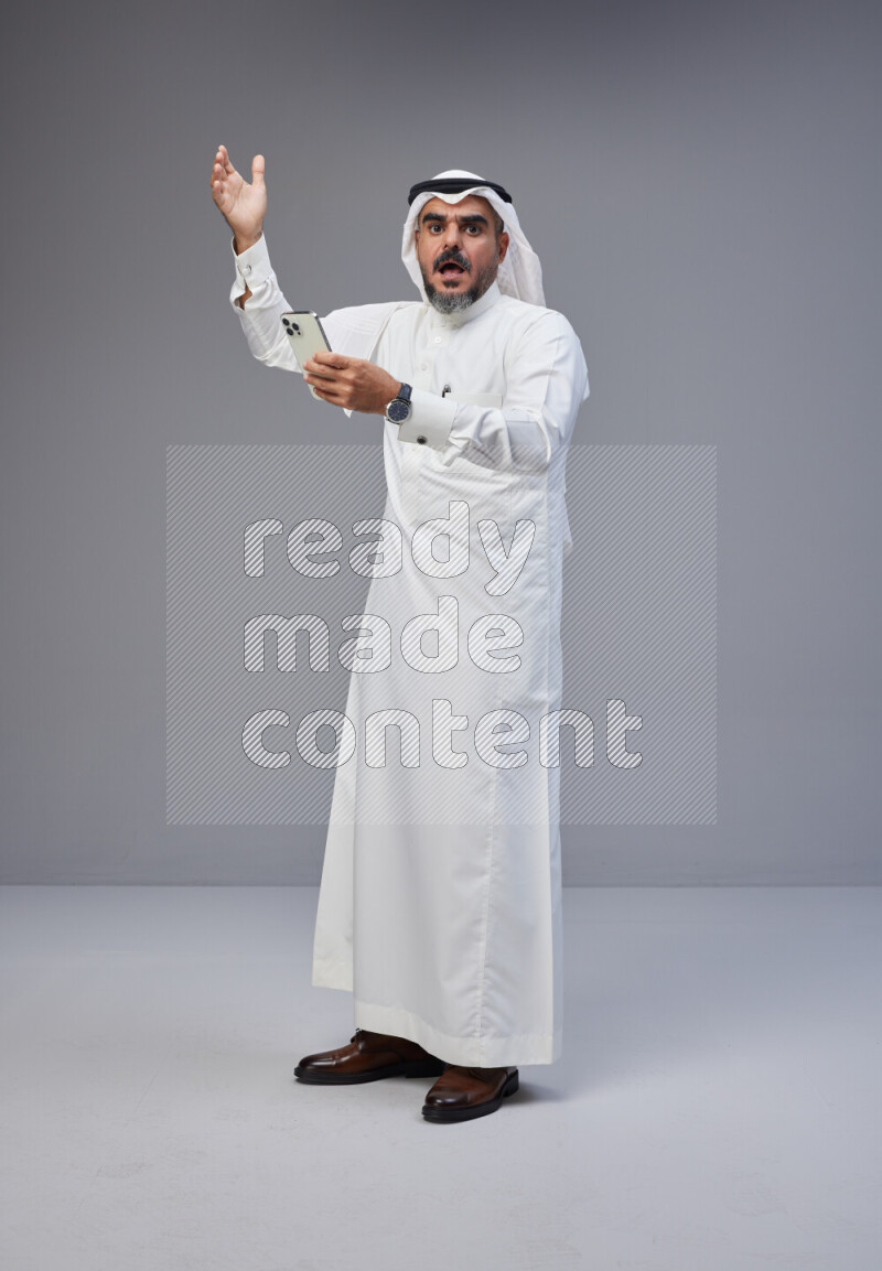 Saudi man Wearing Thob and white Shomag standing texting on phone on Gray background
