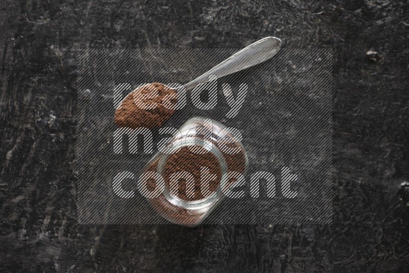A glass spice jar and a metal spoon full of cloves powder on textured black flooring