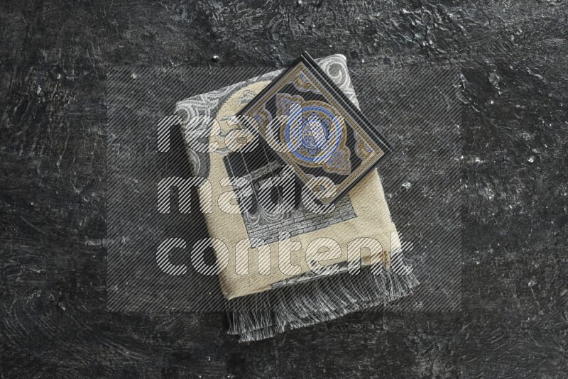 A folded prayer mat with different elements such as quran and prayer beads on black textured background