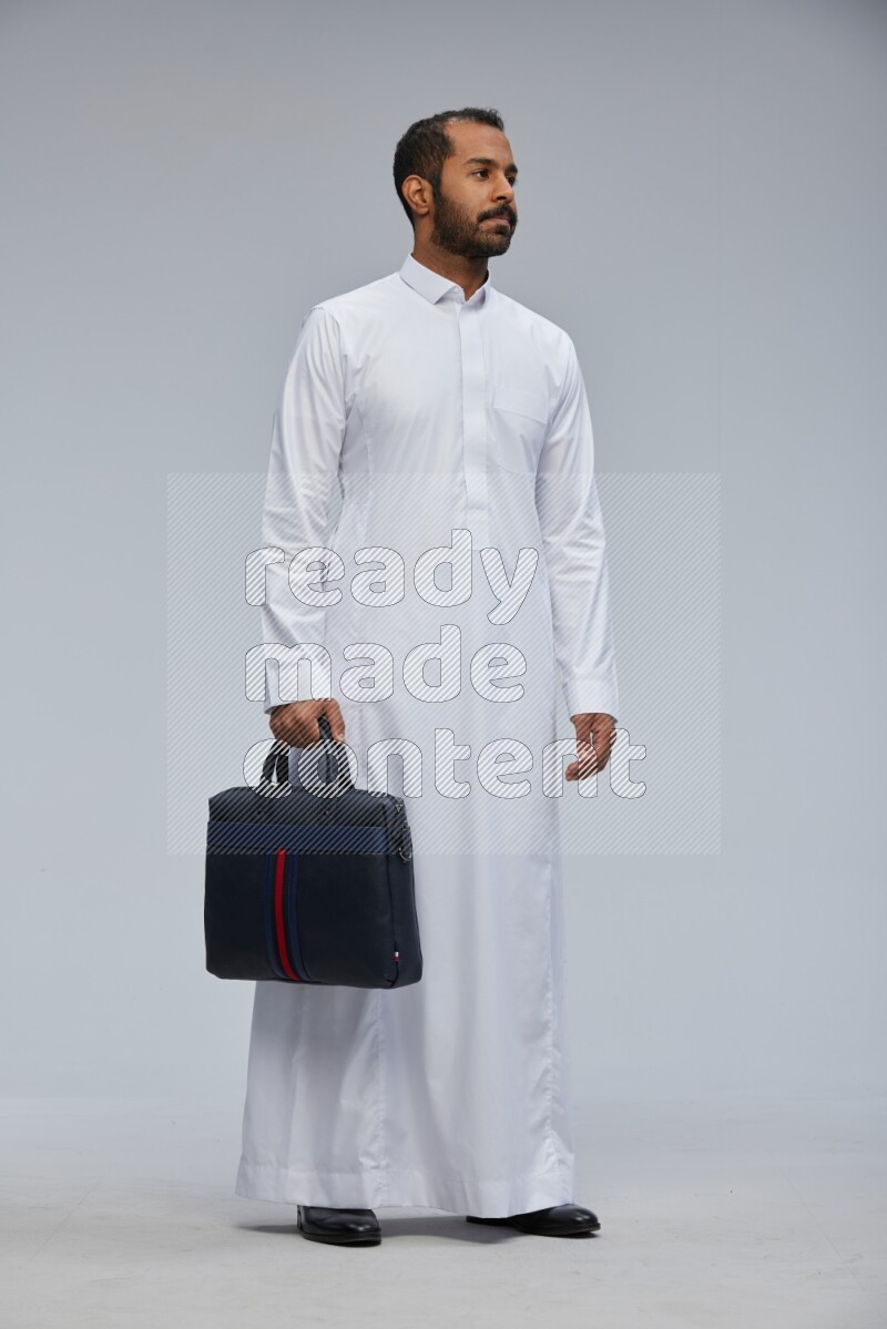 Saudi man wearing thob standing holding suitcase on gray background