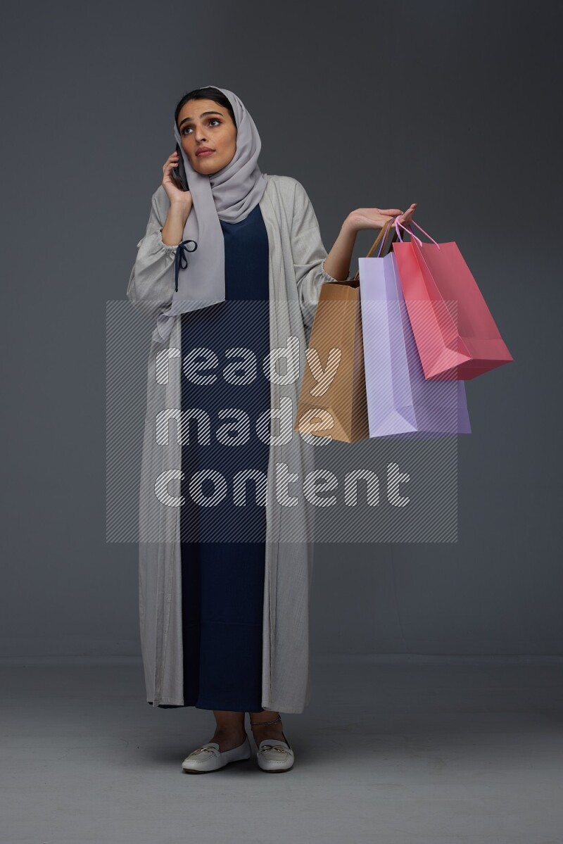 A Saudi woman wearing a light gray Abaya and head scarf standing and holding shopping bags on a grey background