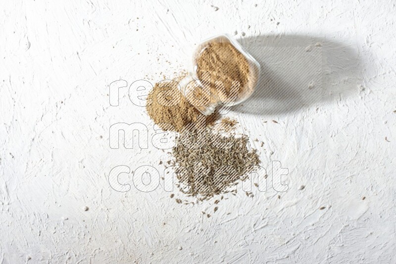 A flipped glass spice jar full of cumin powder and powder spilled out with cumin seeds on textured white flooring