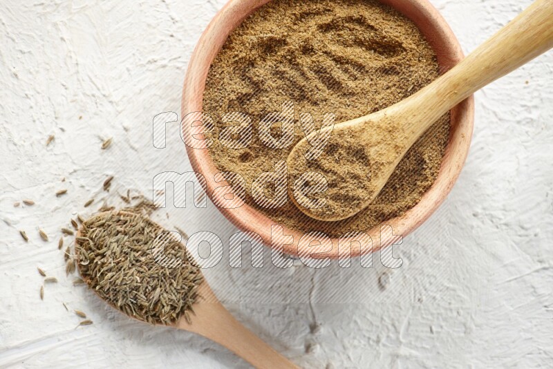 A wooden bowl and 2 wooden spoons full of cumin powder and cumin seeds on textured white flooring
