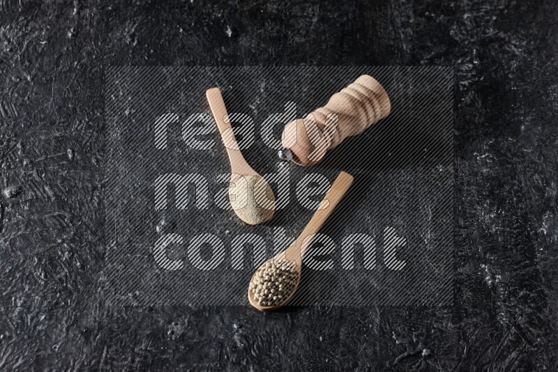 2 wooden spoons one full of white pepper powder and the other with pepper beads and a wooden pepper mill on textured black flooring