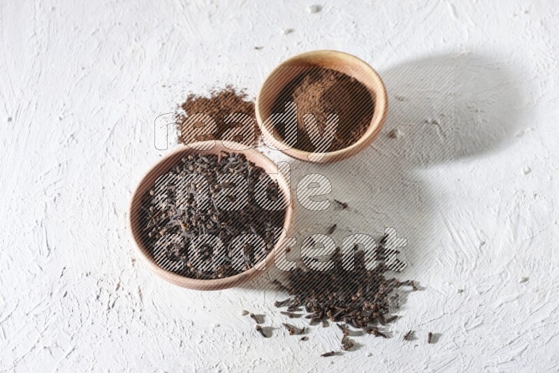 2 wooden bowls full of cloves powder and whole cloves on a textured white flooring