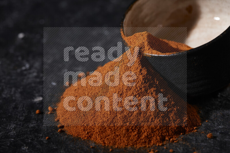 A black pottery bowl full of ground paprika powder with fallen powder from it on black background