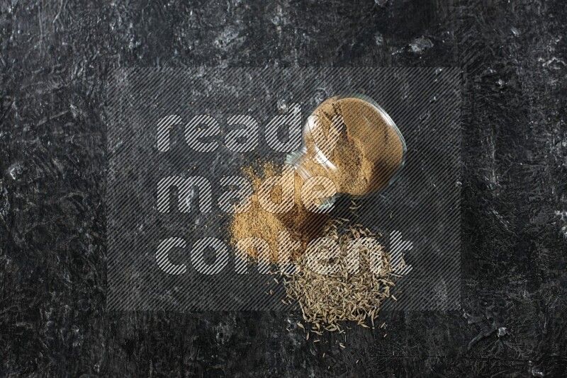 A flipped glass spice jar full of cumin powder with spilled powder and cumin seeds on a textured black flooring