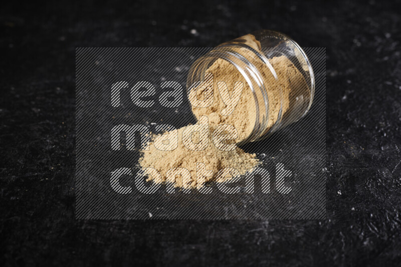 A glass jar full of ground ginger powder flipped with some spilling powder on black background