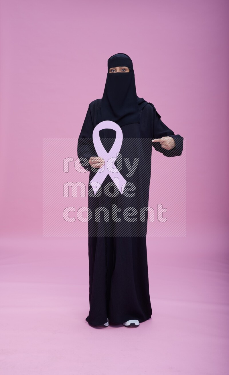Saudi woman wearing abaya and niqab standing holding awareness ribbon on pink background