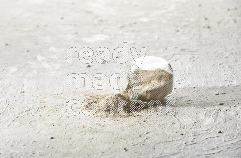 A flipped herbal glass jar full of white pepper powder with spilled powder on textured white flooring