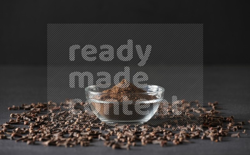 A glass bowl full of cloves powder with cloves grains spread on black flooring