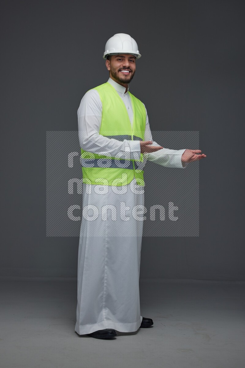 A Saudi man wearing Thobe with a yellow safety vest and white helmet standing and pointing different angles eye level on a gray background