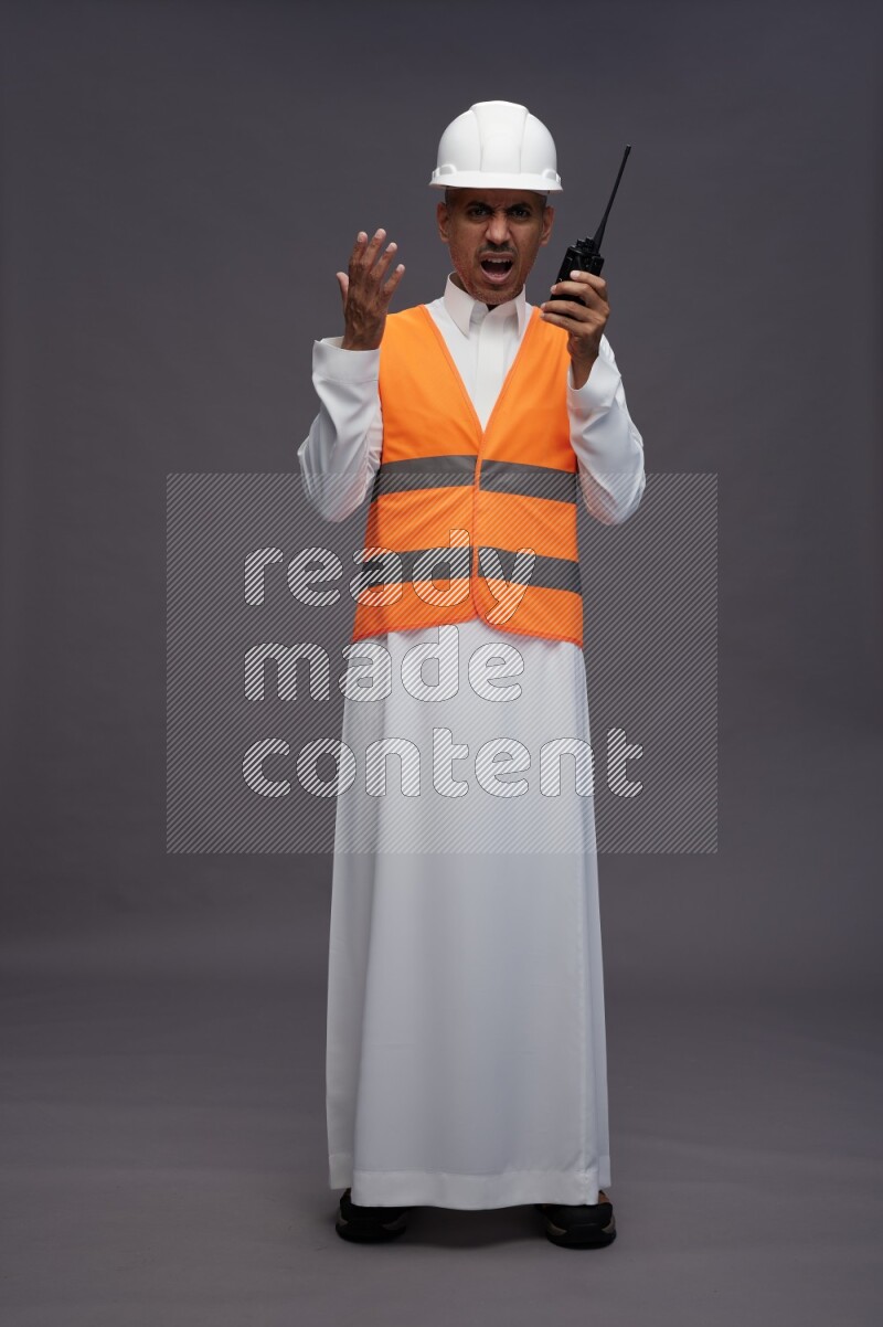 Saudi man wearing thob with engineer vest standing holding walkie-talkie on gray background