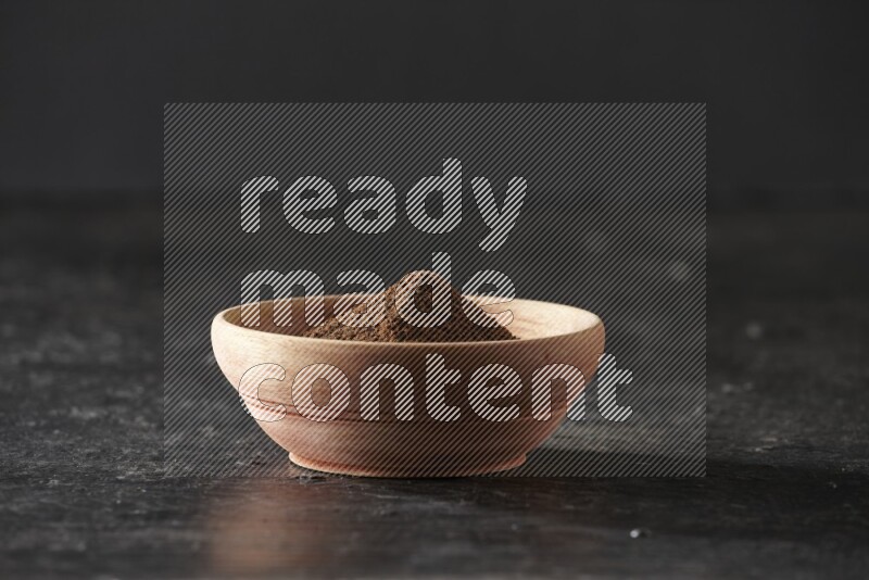 A wooden bowl full of cloves powder on a textured black flooring