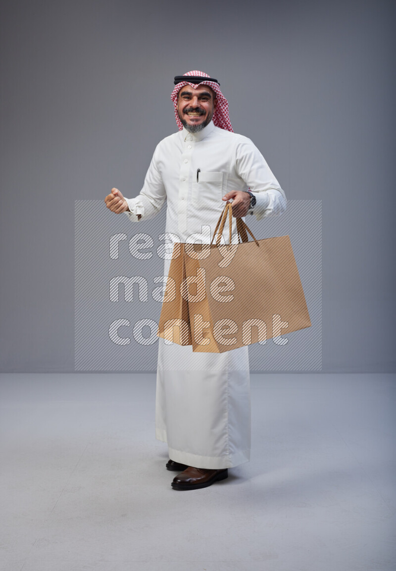 Saudi man Wearing Thob and red Shomag standing holding shopping bag on Gray background