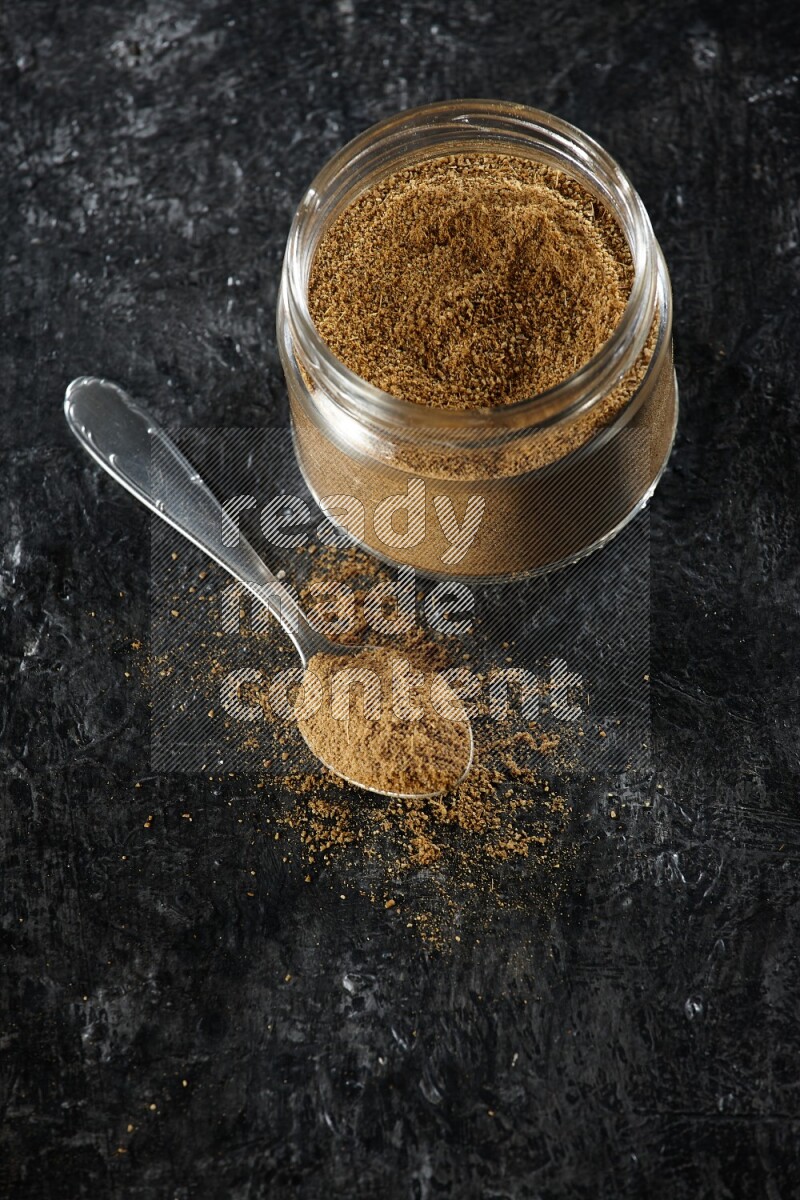 A glass jar and a metal spoon full of cumin powder on a textured black flooring