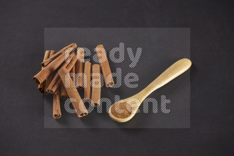 Cinnamon sticks stacked beside a wooden spoon full of cinnamon powder on black background