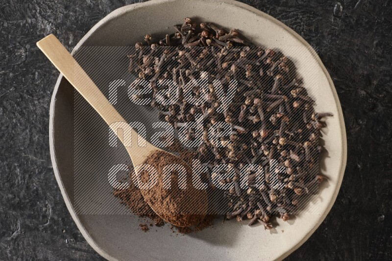 A Pottery plate full of cloves and a wooden spoon full of cloves powder on it on a textured black background