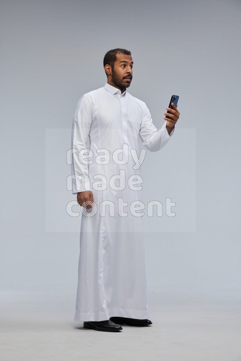 Saudi man wearing Thob standing texting on phone on Gray background
