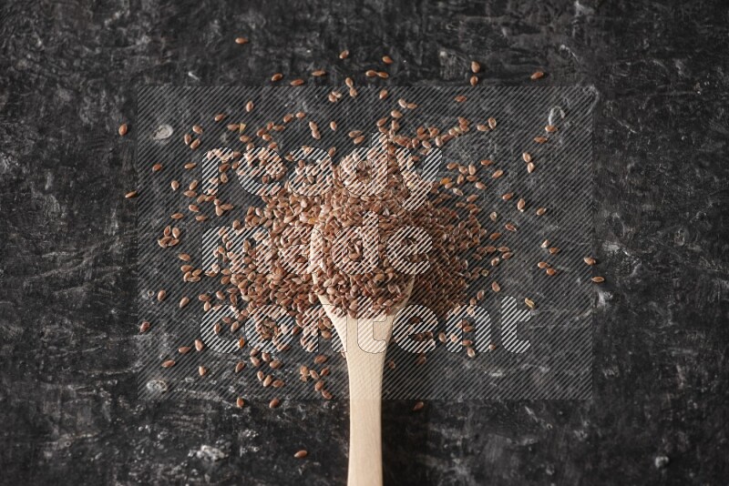 A wooden spoon full of flaxseeds and surrounded by seeds on a textured black flooring