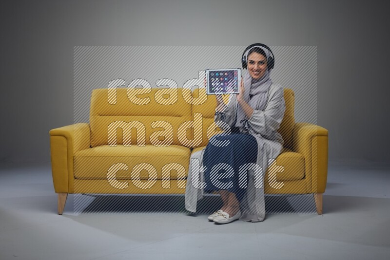 A Saudi female wearing a light gray Abaya and head scarf sitting on a yellow sofa and showing her tablet's screen while wearing headphones eye level on a grey background