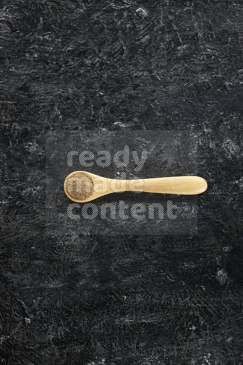 A wooden spoons full of cumin powder on textured black flooring