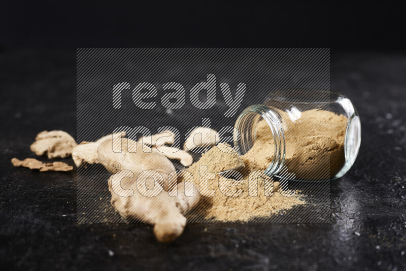 A glass jar full of ground ginger powder flipped with some spilling powder on black background
