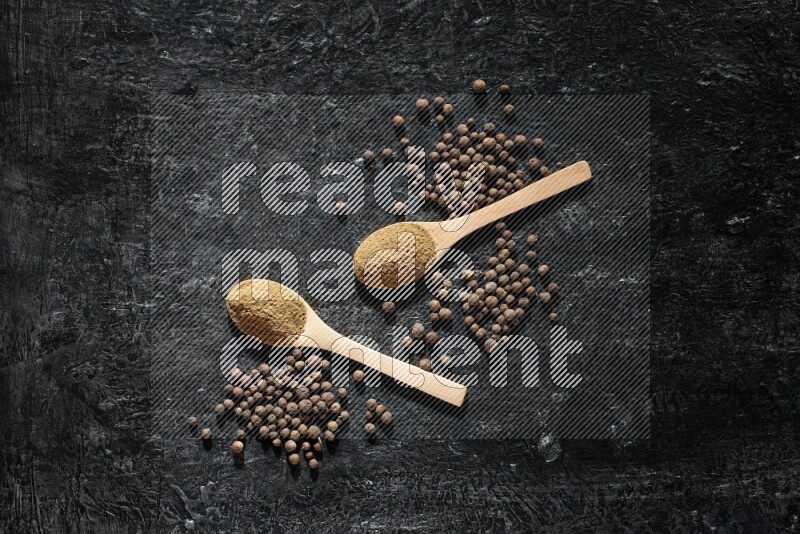 Wooden spoons full of all spice powder and allspice whole balls beside it on a textured black flooring
