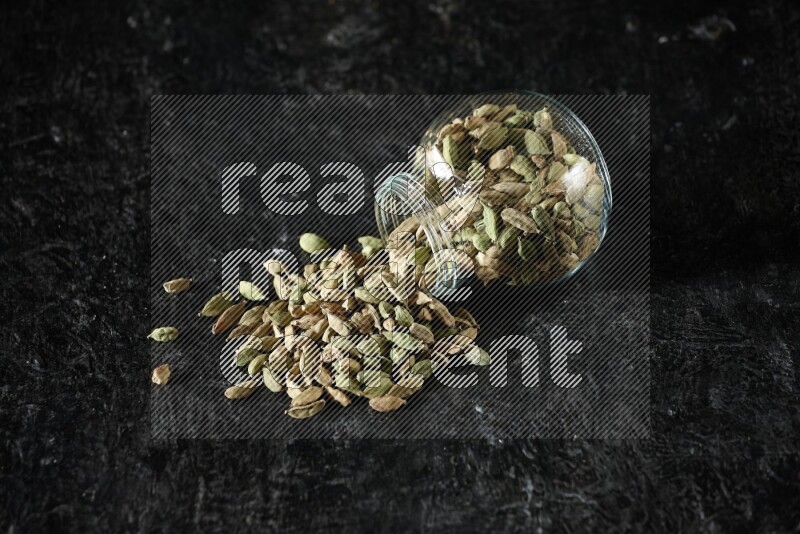 A flipped glass spice jar full of cardamom seeds on textured black flooring