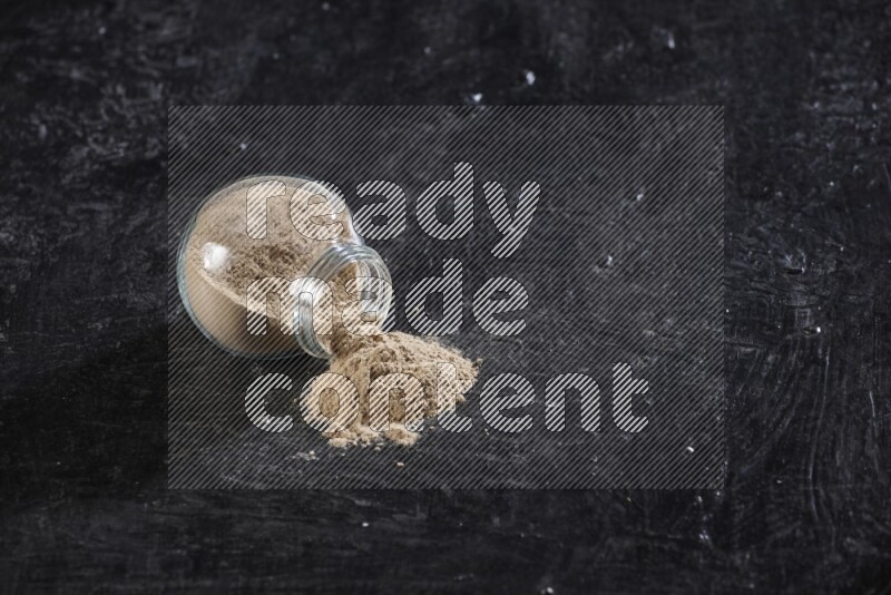 وعاء توابل زجاجي ممتلئ ببودرة الثوم مقلوب والبودرة متناثرة منه علي خلفية سوداء