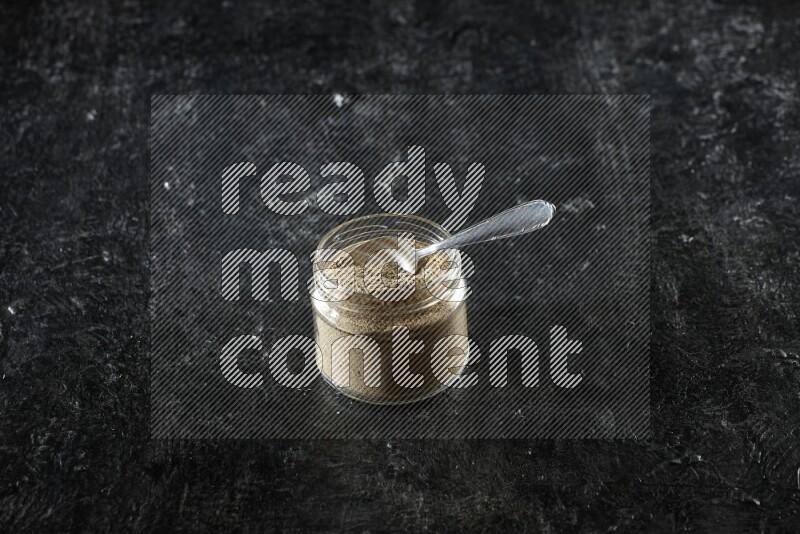 A glass jar and metal spoon full of cardamom powder on textured black flooring