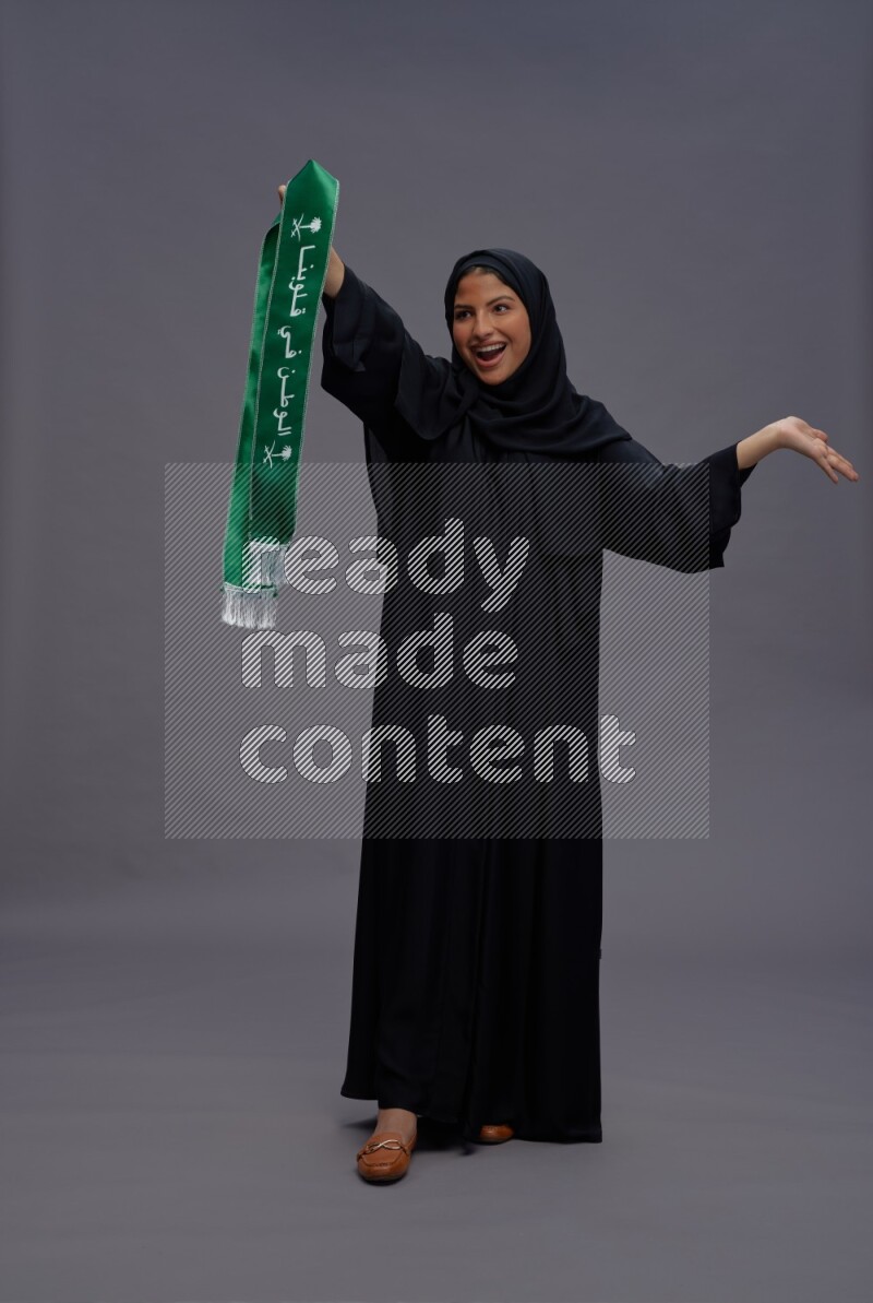Saudi woman wearing Abaya standing holding Saudi flag on gray background