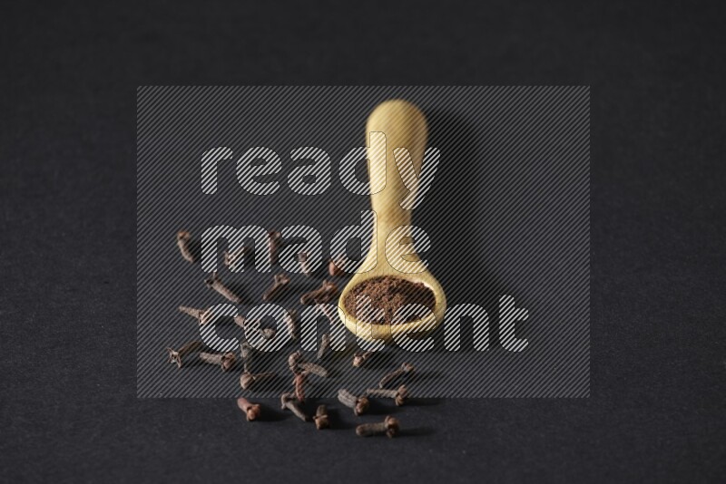 ملعقة خشبية ممتلئة ببودرة القرنفل مع حبوب القرنفل المتناثرة على خلفية سوداء